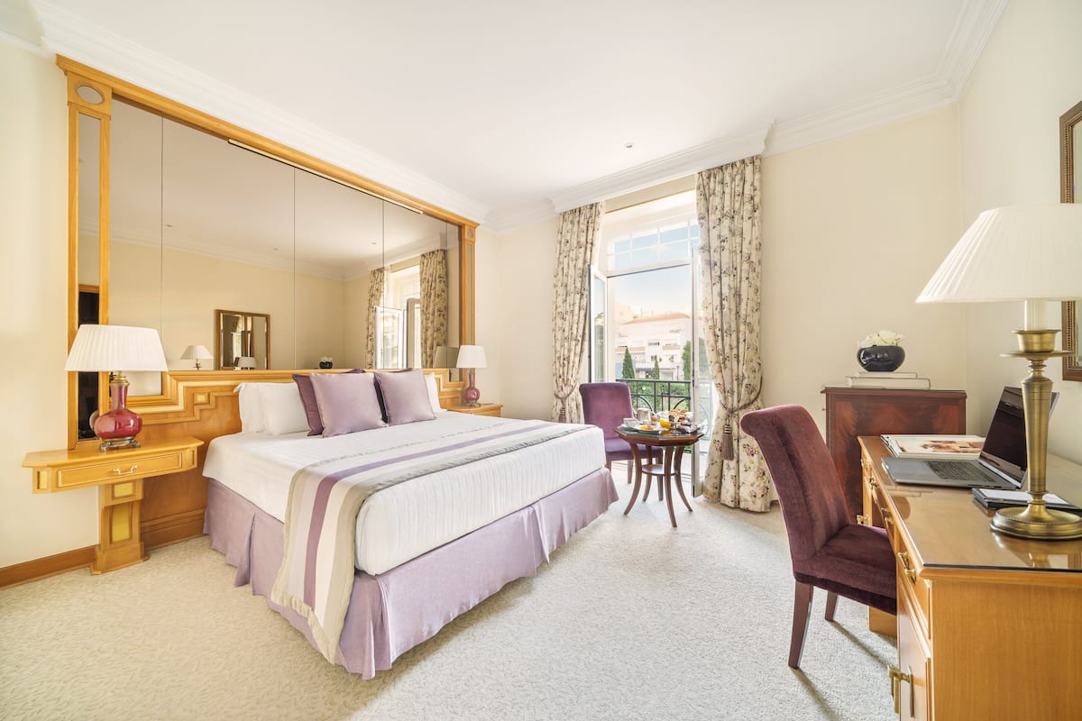 A hotel room with a large bed, a desk with a laptop, a chair, a small table with a lamp, and a large mirror.