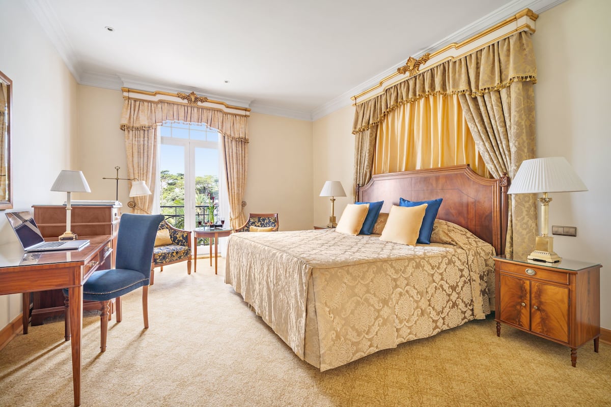 A hotel room with a large bed, a desk with a laptop, a chair, and a small table with a lamp.