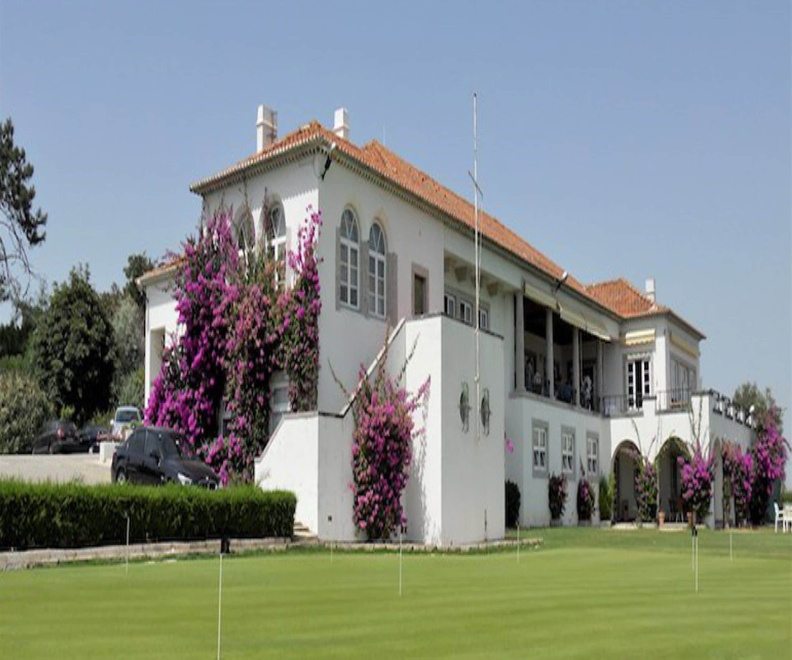 Un edificio grande y blanco con techo de tejas rojas, rodeado de setos cuidados y vibrantes flores de buganvilla rosadas.