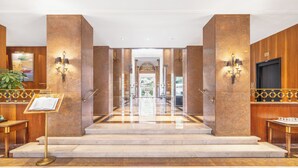 A hotel lobby with marble flooring, a reception desk, a staircase leading to the upper floor, and wall-mounted lighting. - Palácio Estoril Hotel, Golf & Wellness (Cascais)