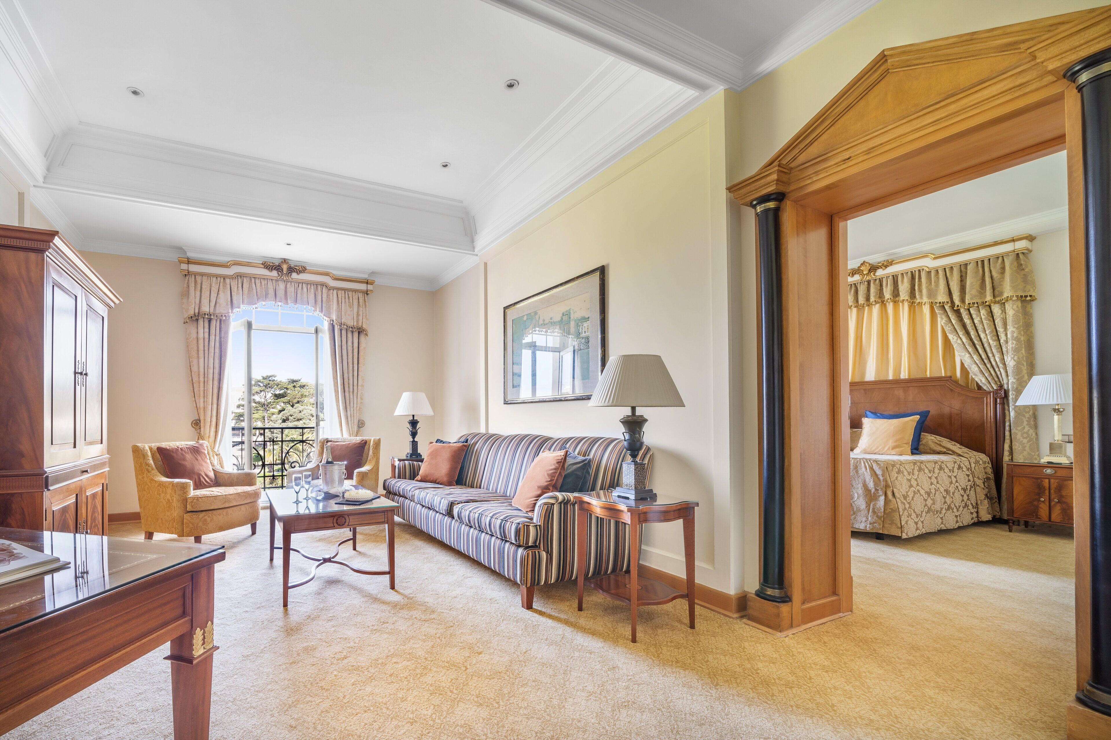 Uma sala de estar ampla, com um sofá, poltronas e uma mesa de centro. Há uma janela grande com cortinas, um quadro na parede e vista para o exterior.