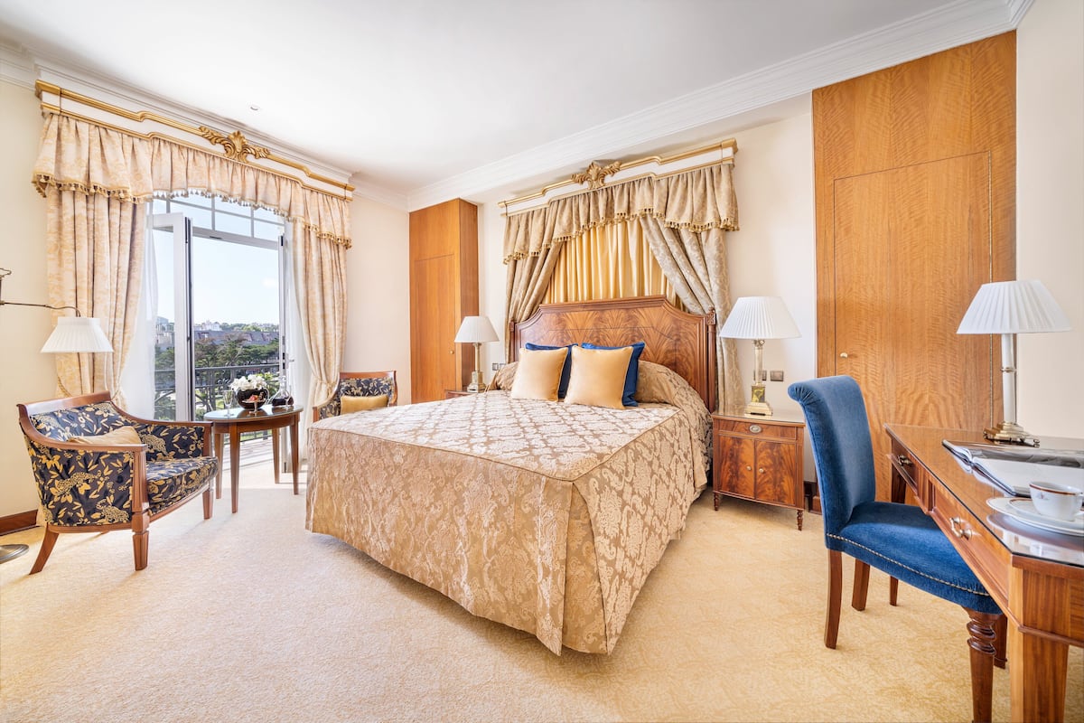 A spacious hotel room with a large bed, a sitting area with a chair and a table, and a desk with a lamp.