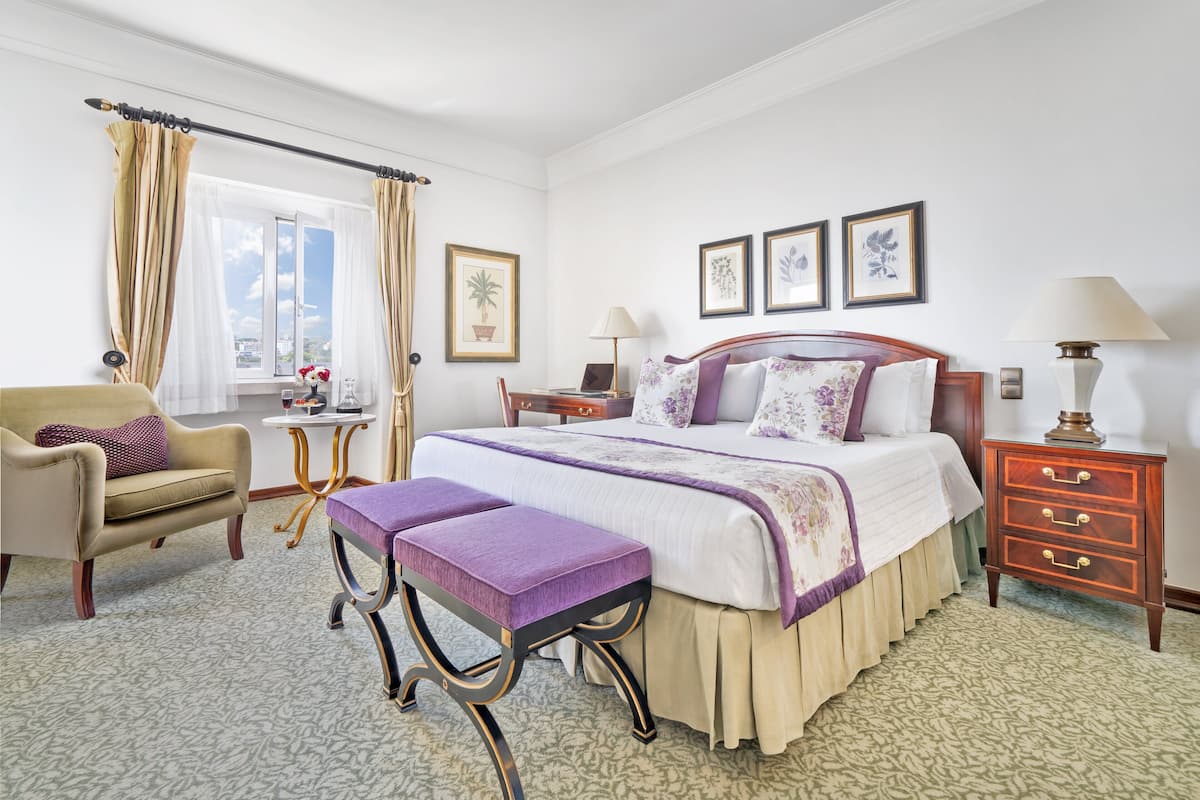 A hotel room with a bed, a chair, a small table, a bench, and a nightstand with lamps.
