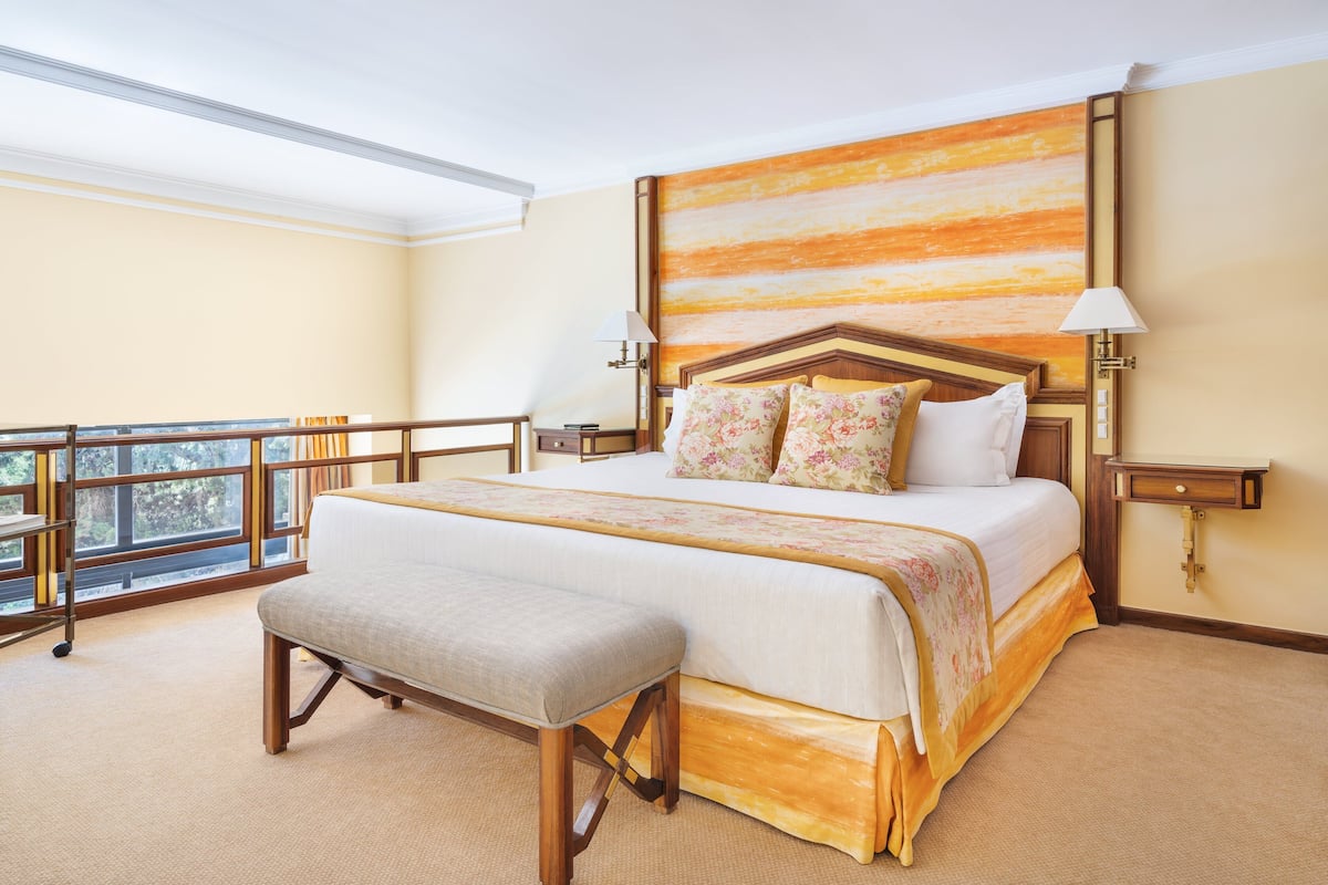 A spacious hotel room with a large bed, a bench, a view of the outdoors, and a balcony.