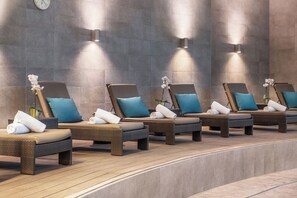 Une rangée de chaises longues avec des coussins et des serviettes, adossées à un mur orné d’une horloge et de lampes fixées au mur.