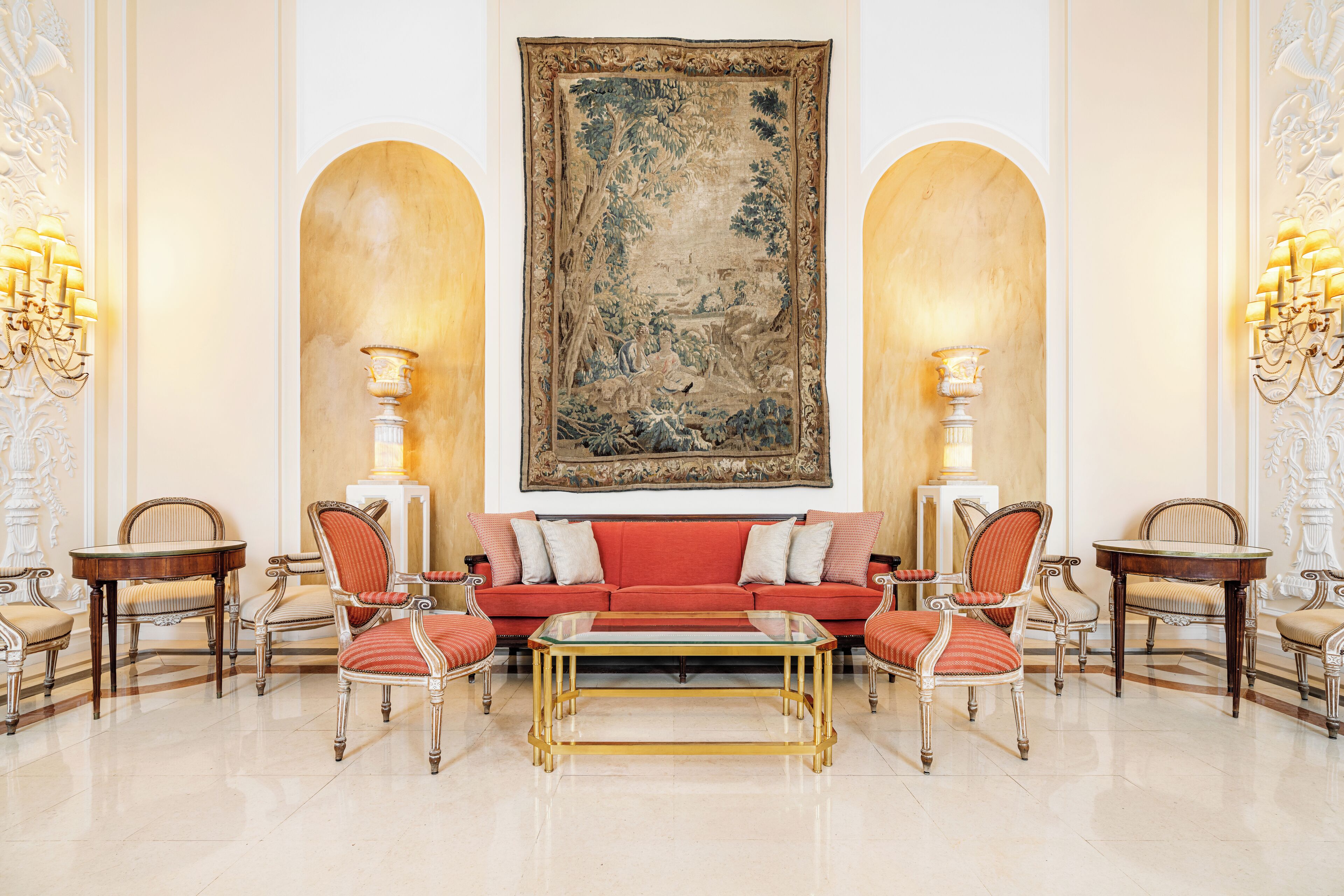 Un lobby lujoso con un sofá rojo, sillas a juego, una mesa de centro de cristal, apliques de pared ornamentados y un gran tapiz.