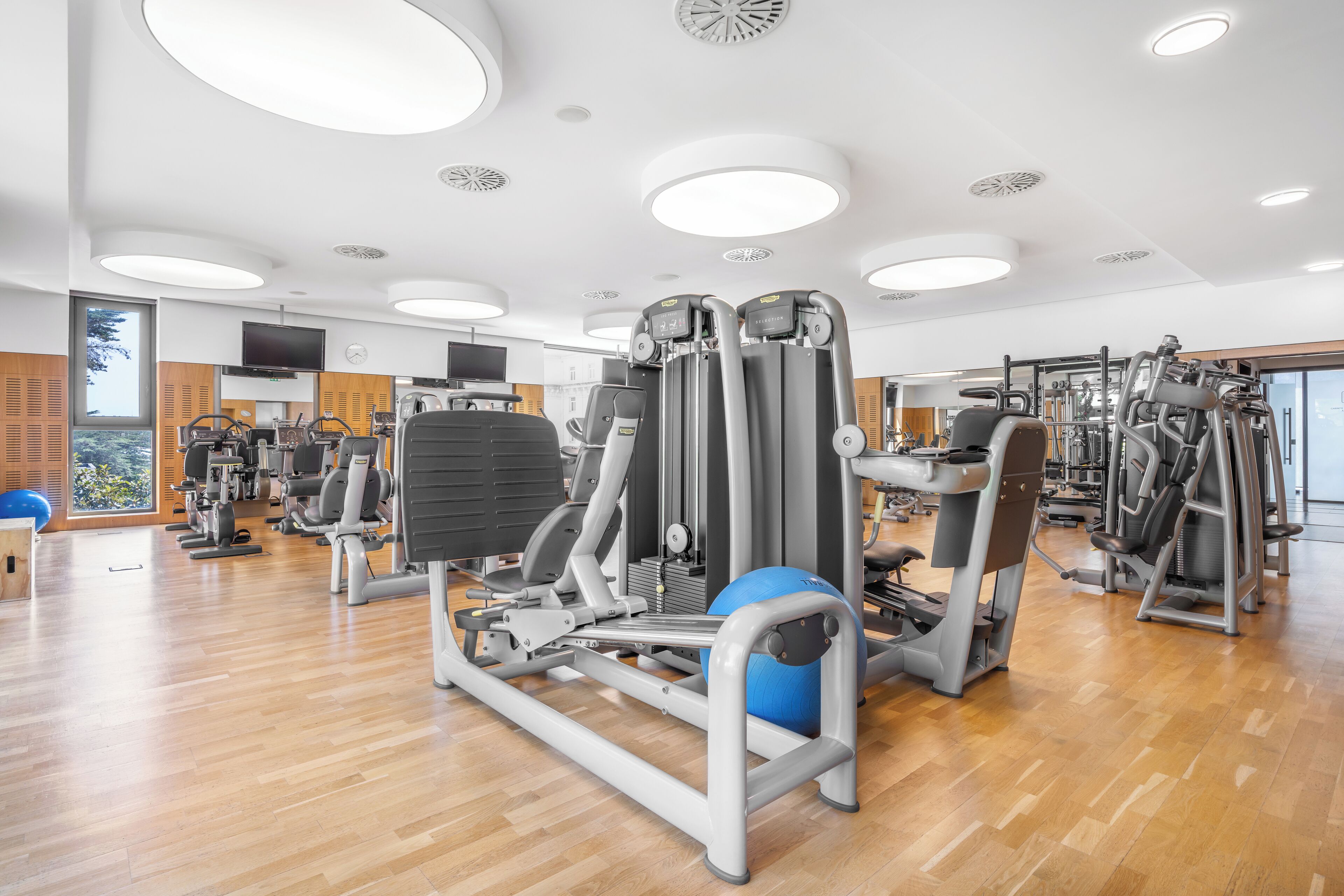 A modern gym with various exercise machines, a television, and a large window providing natural light.