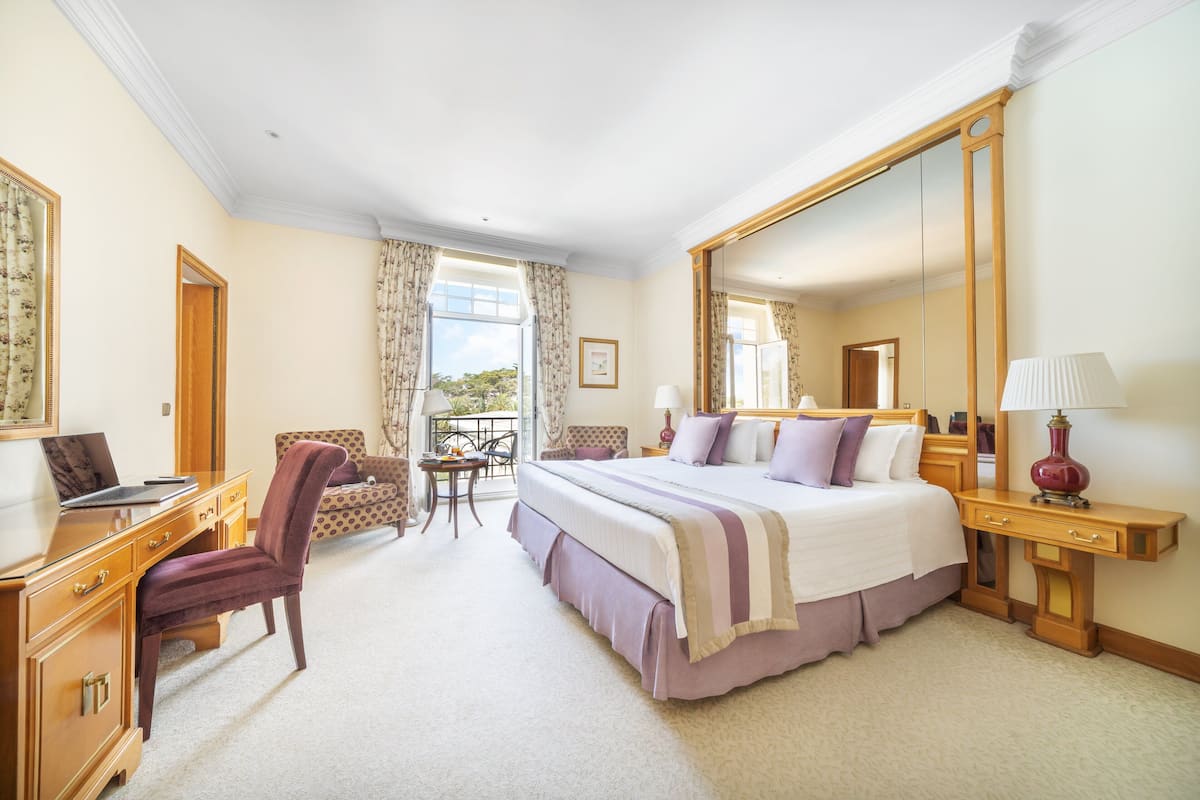 A hotel room with a large bed, a desk with a laptop, a chair, a small table, and a balcony with a view.