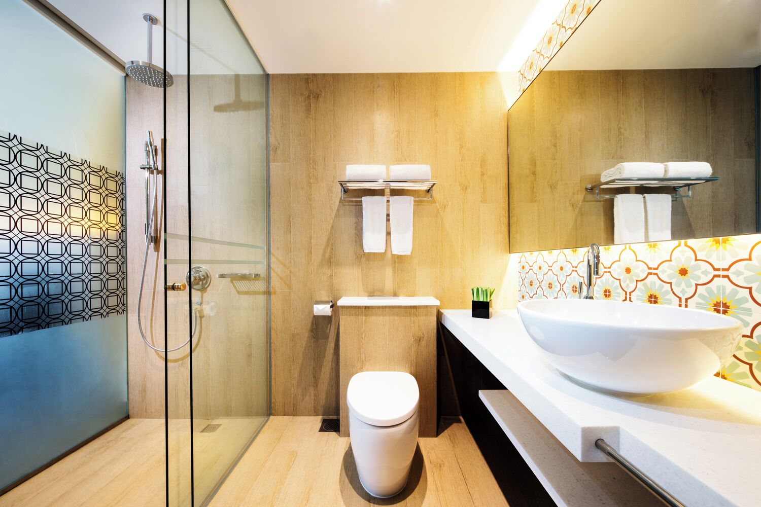 Un baño moderno con cabina de ducha de vidrio, inodoro, lavabo de gran tamaño y un salpicadero de azulejos estampados.
