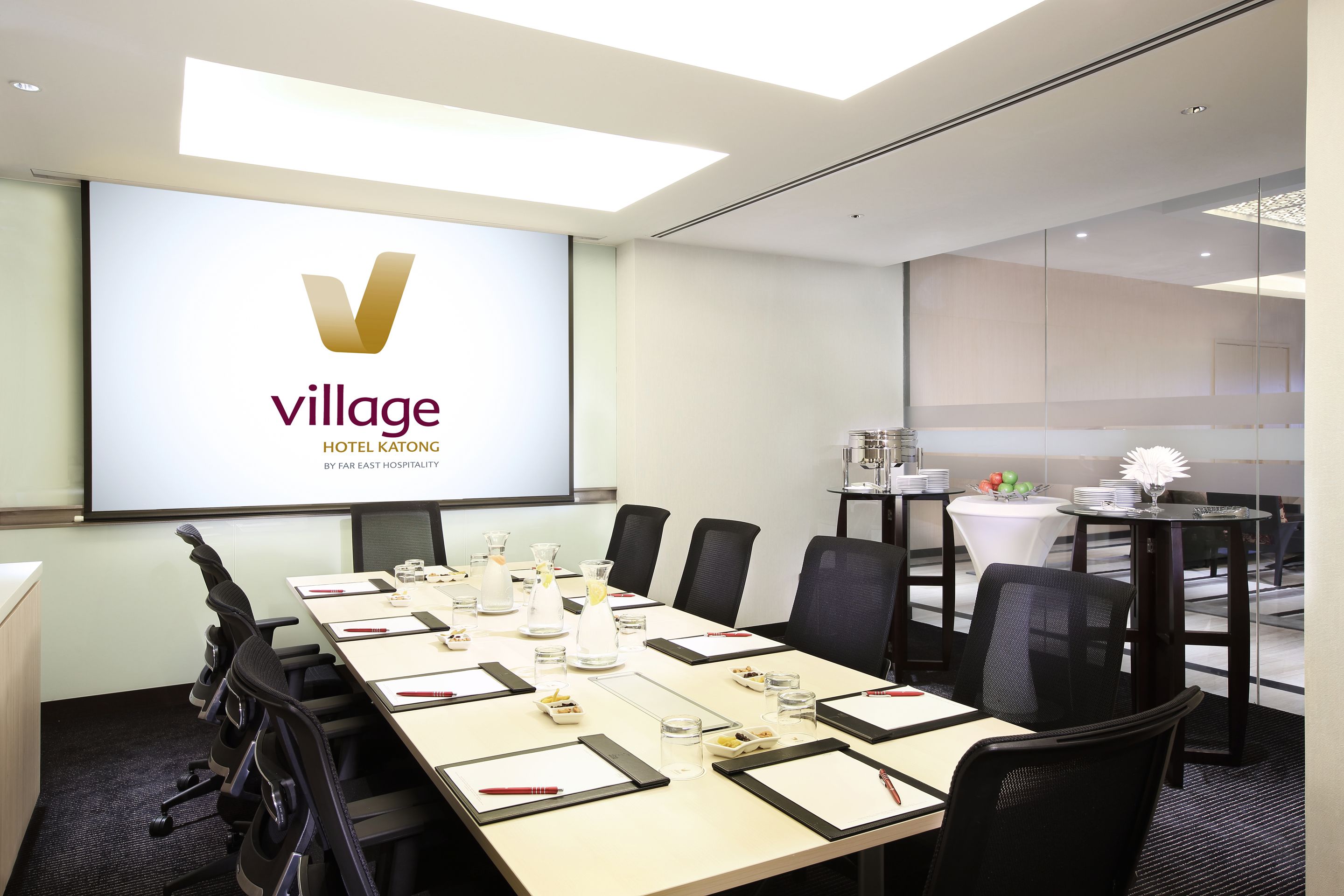 Una sala de conferencias con una larga mesa, sillas negras y una pantalla que muestra el logotipo de Village Hotel Katong by Far East Hospitality.