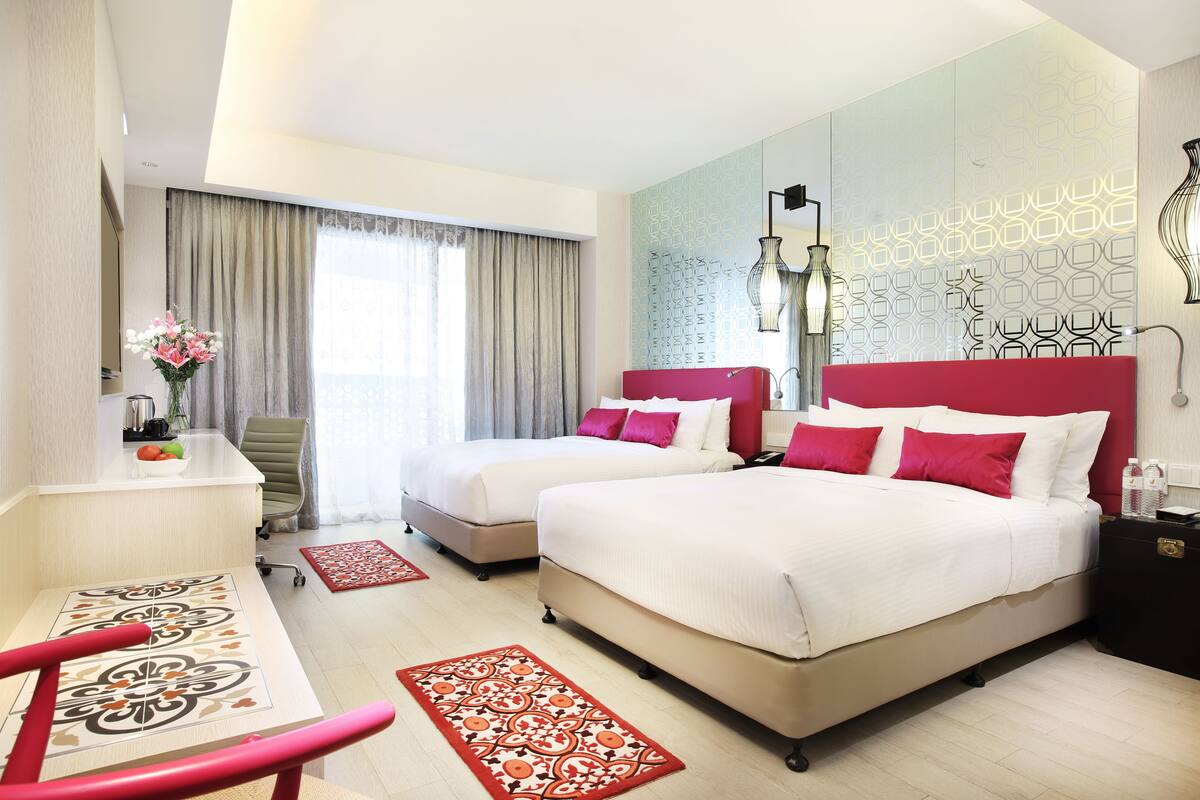Habitación de hotel con dos camas, un escritorio, una silla, un tapete con estampado rojo y blanco, y un panel decorativo en la pared.