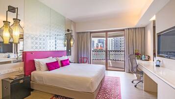 A hotel room with a bed, a desk, a TV, a bathroom, and a balcony with a view of buildings.