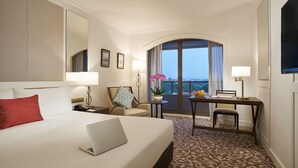 Een hotelkamer met een groot bed, een bureau met een laptop, een stoel, een kleine tafel met een plant en een groot raam met uitzicht.