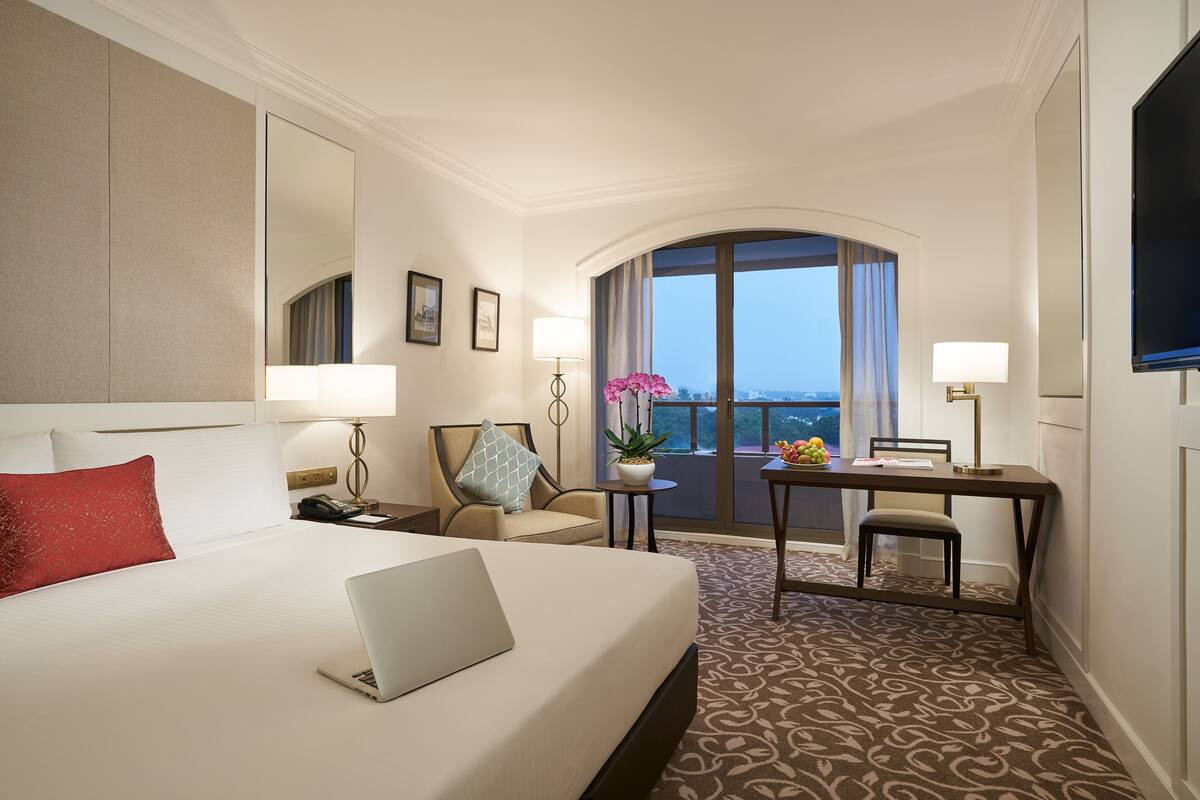 Une chambre d’hôtel avec un grand lit, un bureau avec un ordinateur portable, une chaise, une petite table avec une plante et une grande fenêtre donnant sur un beau paysage.