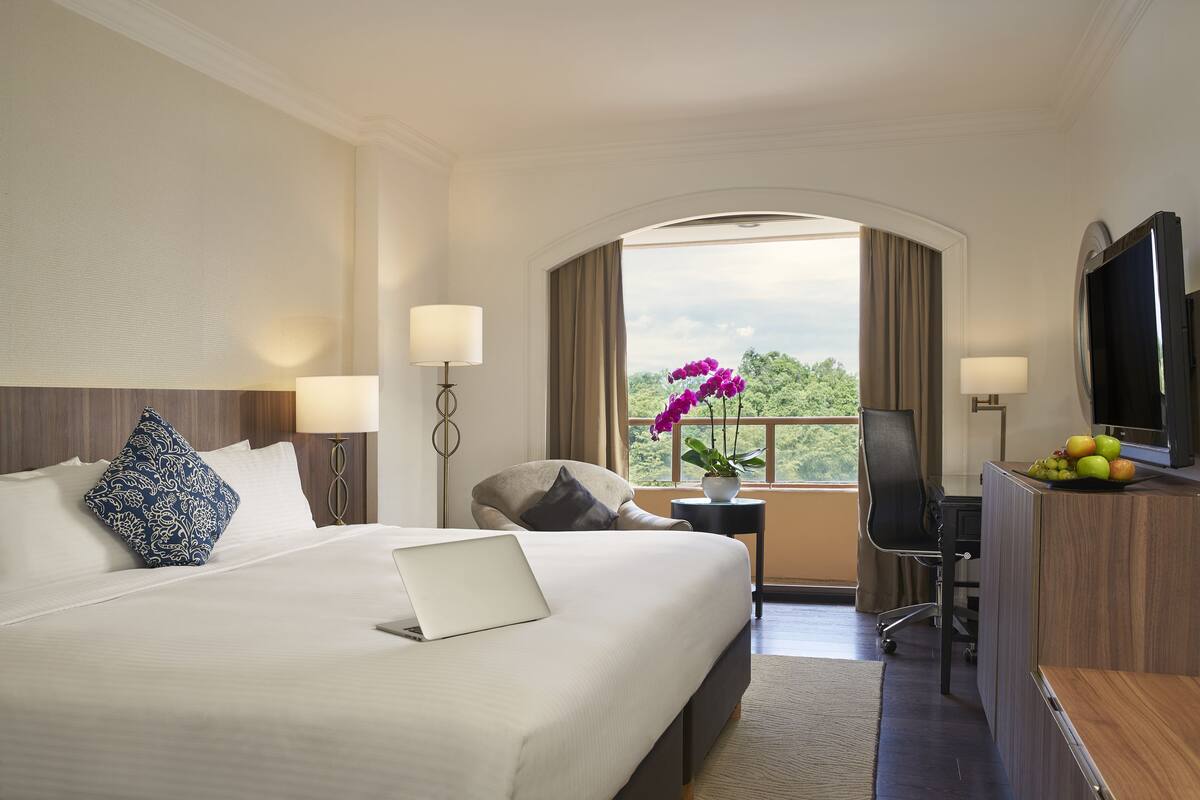 Une chambre d’hôtel avec un grand lit, un bureau avec un ordinateur portable, une télévision et une vue sur la verdure environnante.