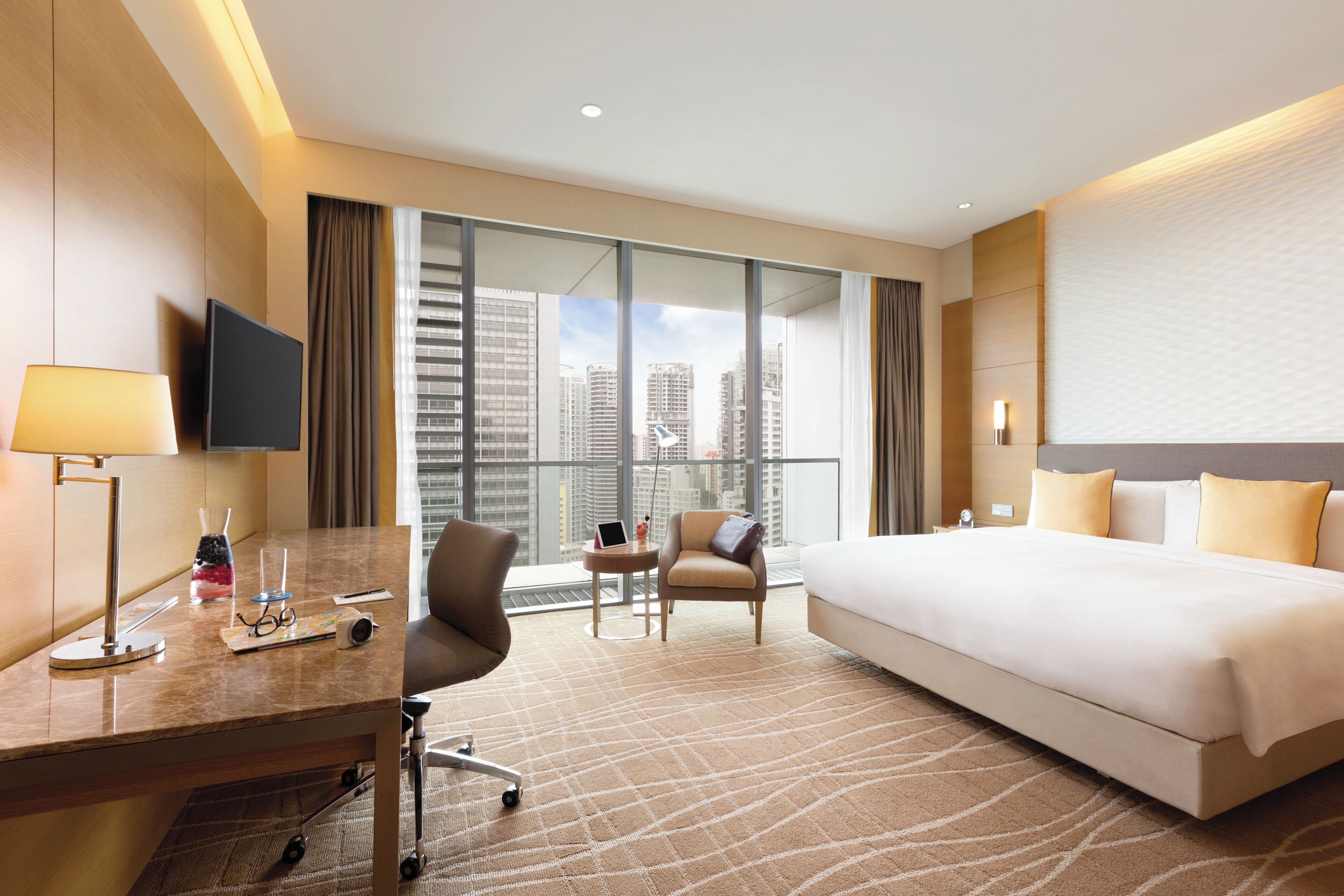 A hotel room with a large bed, a desk with a lamp, a chair, a small table with a laptop, and a view of tall buildings through large windows.