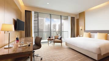 A hotel room with a large bed, a desk with a lamp, a chair, a small table with a laptop, and a view of tall buildings through large windows.