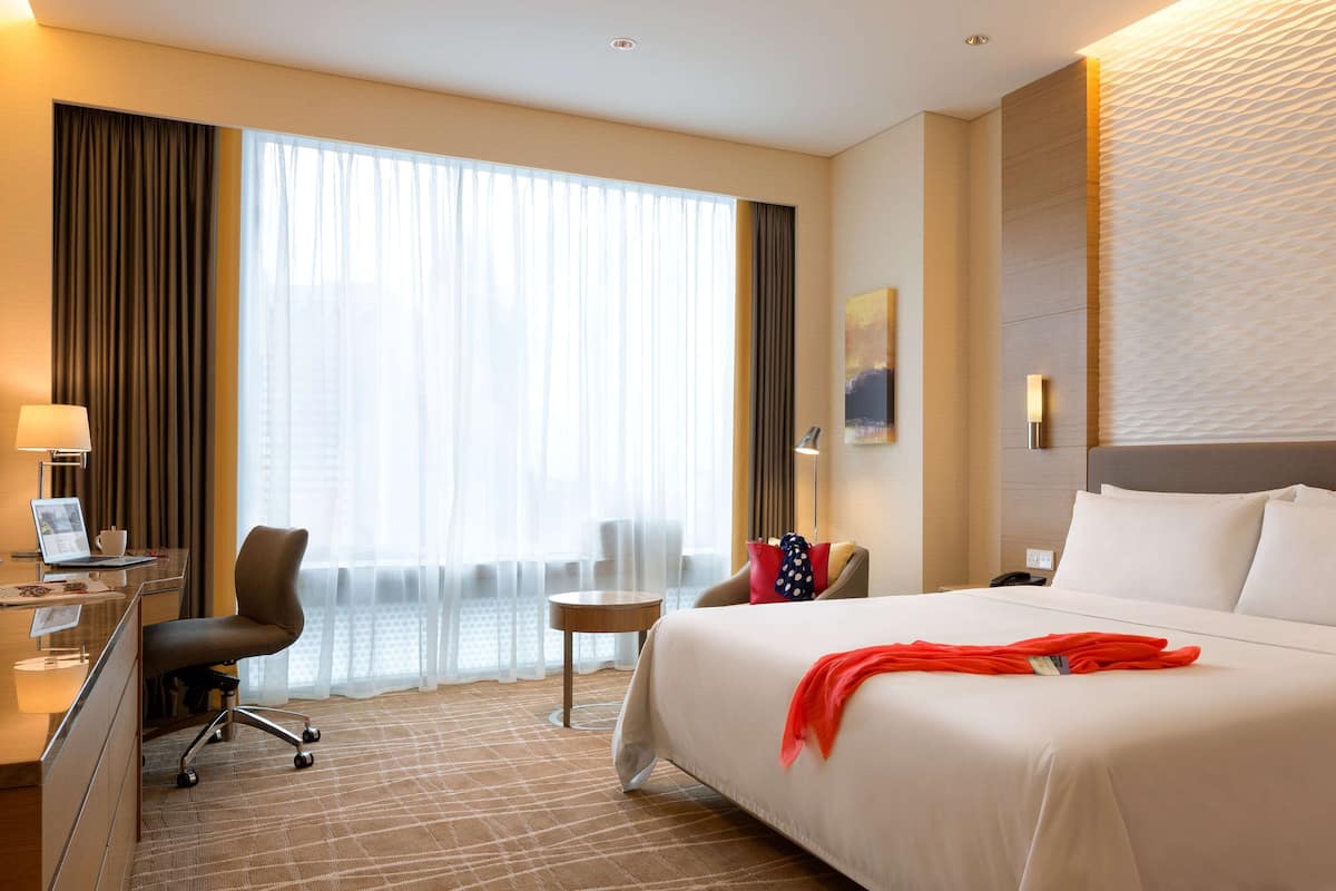 Habitación de hotel con una cama grande, un escritorio con una computadora portátil, una silla, una mesita y una ventana con cortinas.