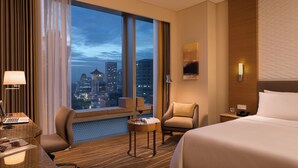 Camera d'albergo con un letto grande, una scrivania con un portatile, una sedia, un tavolino con una bevanda e vista sulla città attraverso ampie finestre.