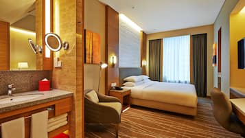 A hotel room with a bed, a desk, a chair, a nightstand, a mirror, a bathroom with a sink and towel rack.