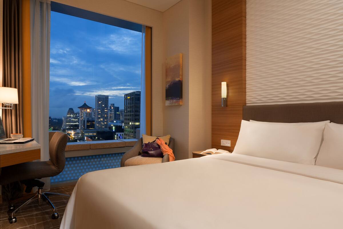 Habitación de hotel con un ventanal grande que ofrece vistas a la ciudad, una cama con ropa blanca, un escritorio con lámpara y una silla.