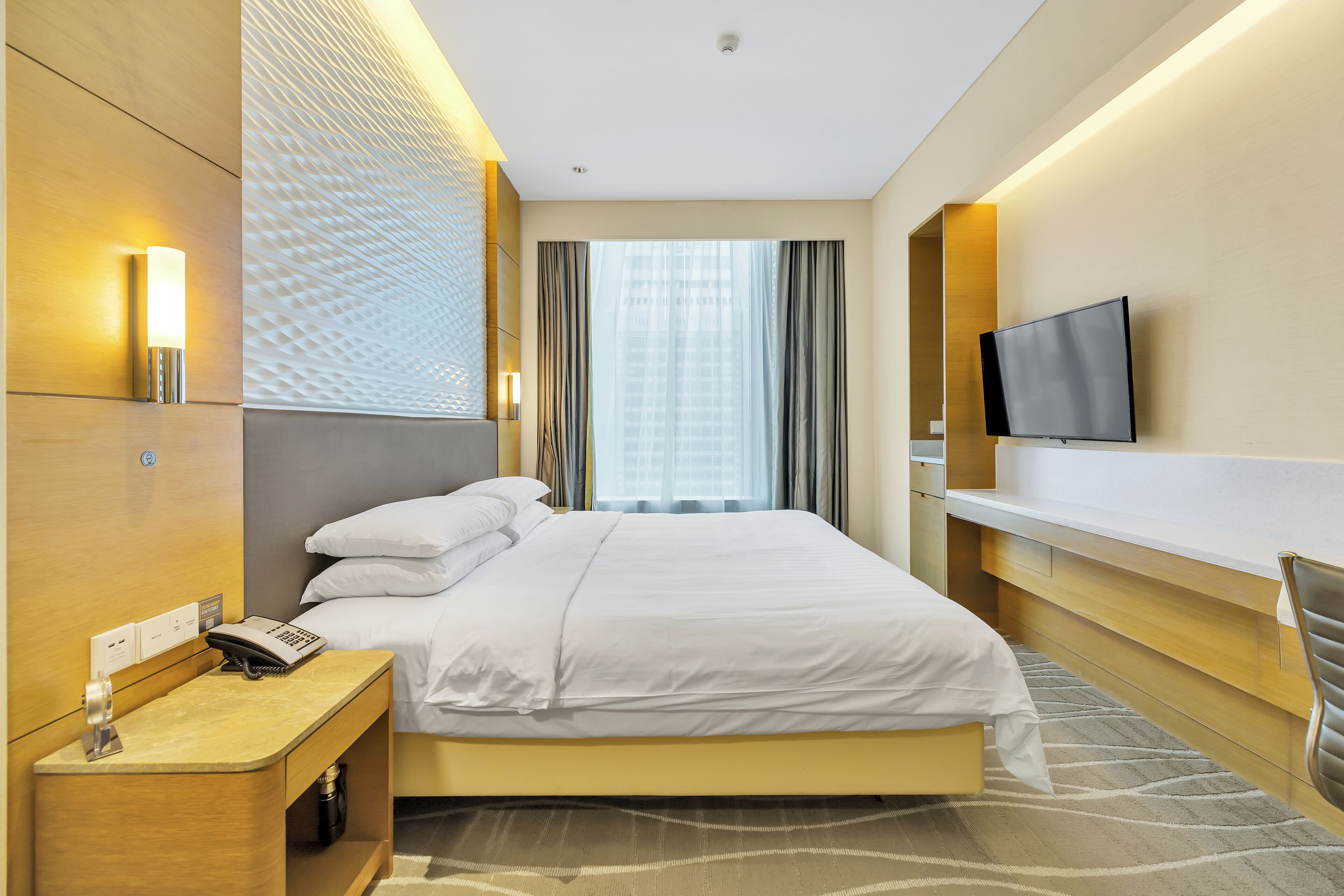 A modern hotel room with a large bed, bedside table with a phone, a flat-screen TV mounted on the wall, and a window with curtains.