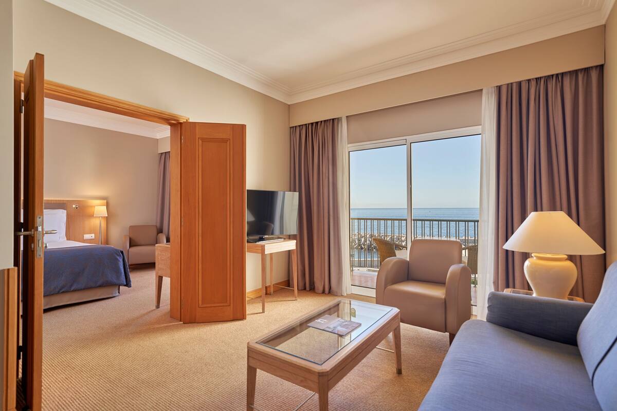 Ein Hotelzimmer mit Bett, Schreibtisch mit Computer, Sofa, Couchtisch und einem Balkon mit Meerblick.
