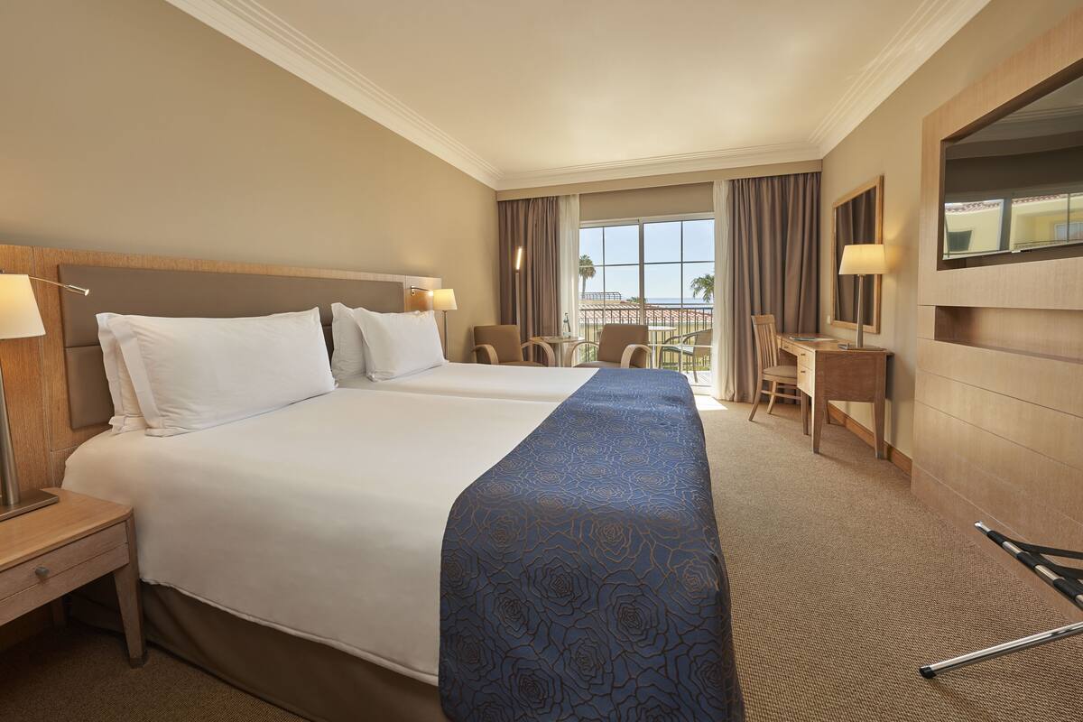 Ein Hotelzimmer mit zwei Betten, einem Schreibtisch, Stühlen, einem Fernseher und einem Balkon mit Blick.