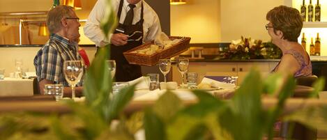 Un interior de restaurante con un camarero sirviendo una cesta de pan a una pareja sentada.