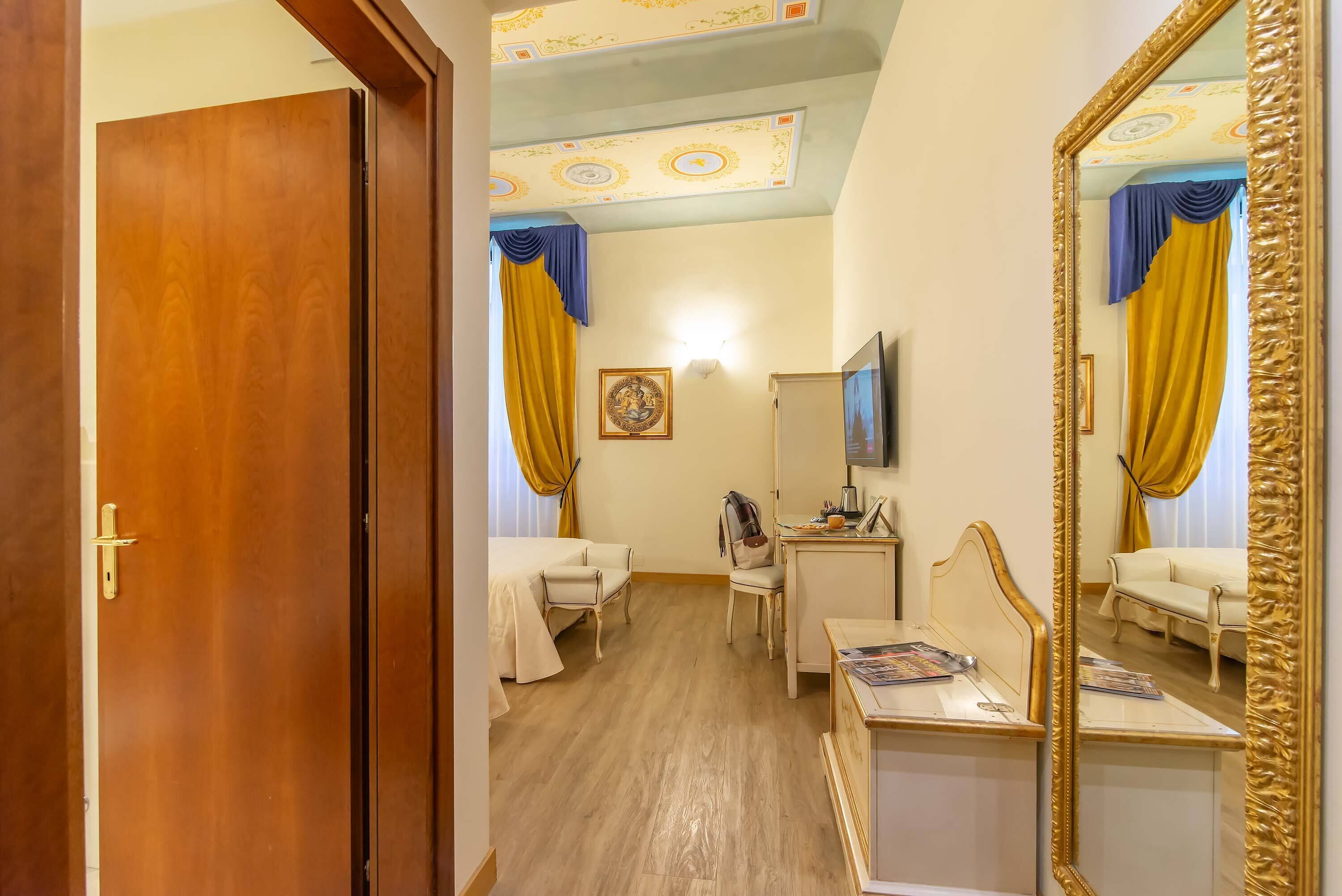 Habitación de hotel con puerta de madera, una cama, un escritorio, una silla, televisor y un espejo.