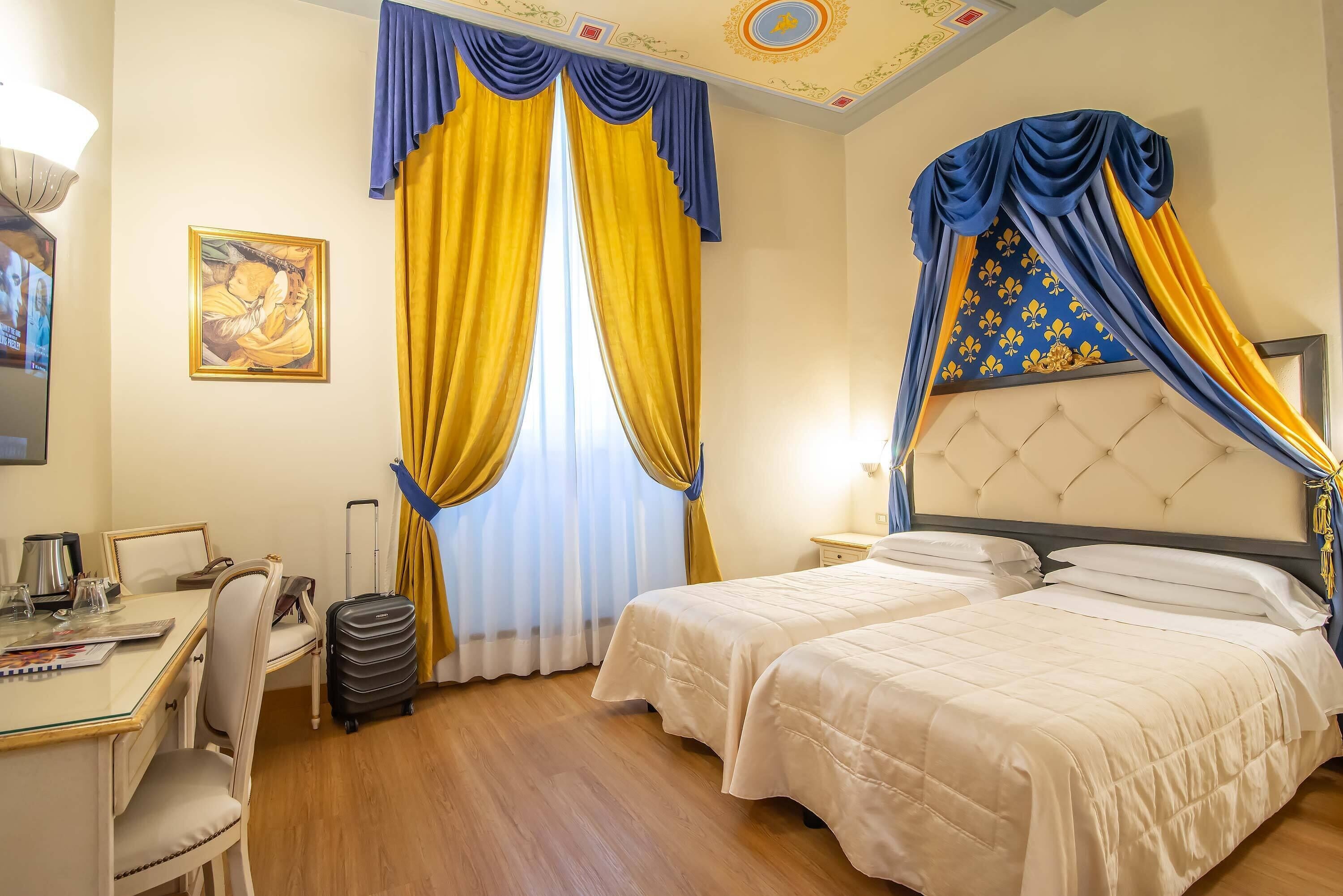 A hotel room with two beds, a desk, a chair, a suitcase, and a painting on the wall.