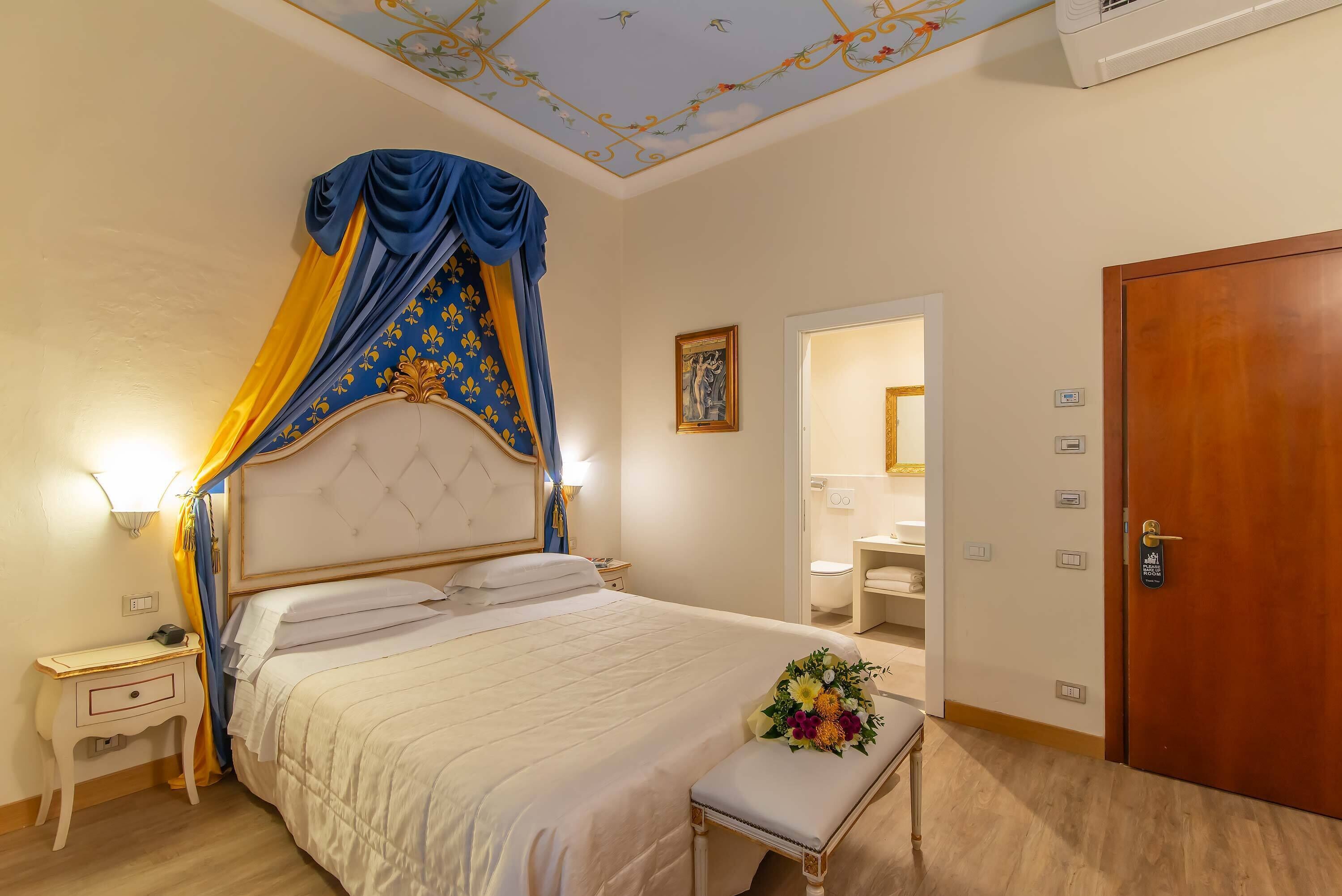 A neatly arranged bedroom with a large bed, a bedside table, a bench with a floral arrangement, and a wooden door.