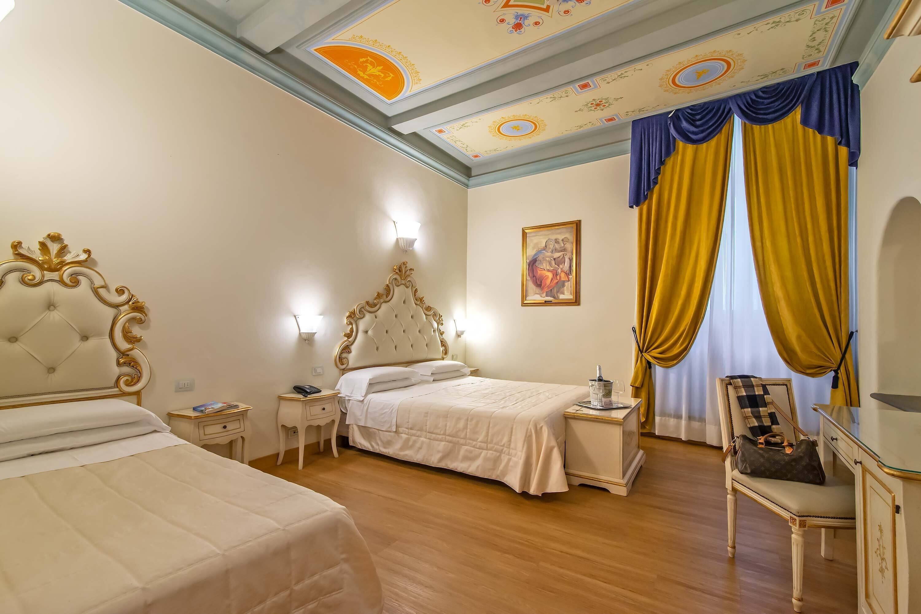 Habitación de hotel con una cama grande, una cama más pequeña, una silla, una mesita de noche, una lámpara y un cuadro en la pared.
