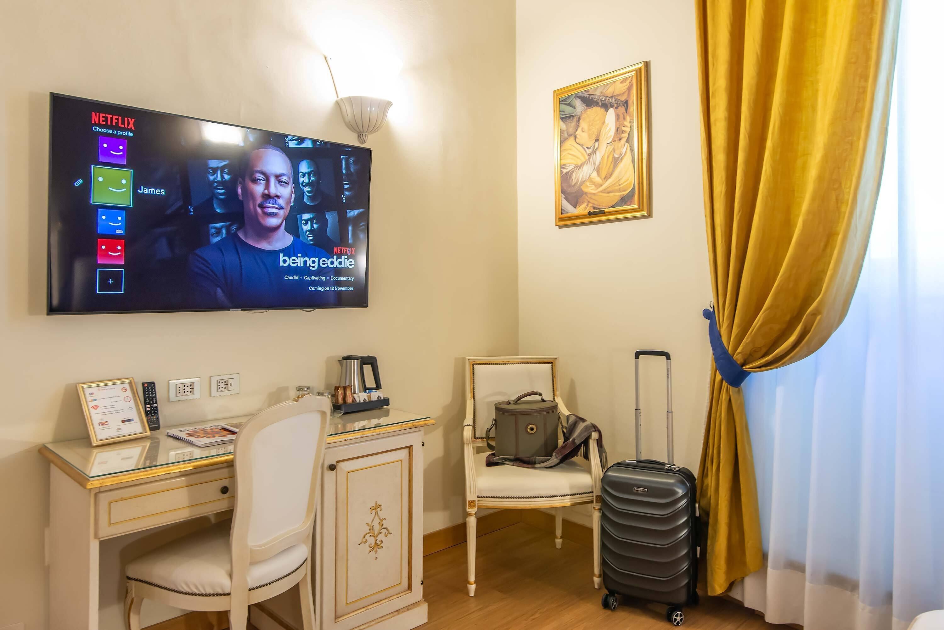 A hotel room with a desk, chair, a TV displaying a Netflix interface, a framed certificate, a suitcase, and a painting on the wall.