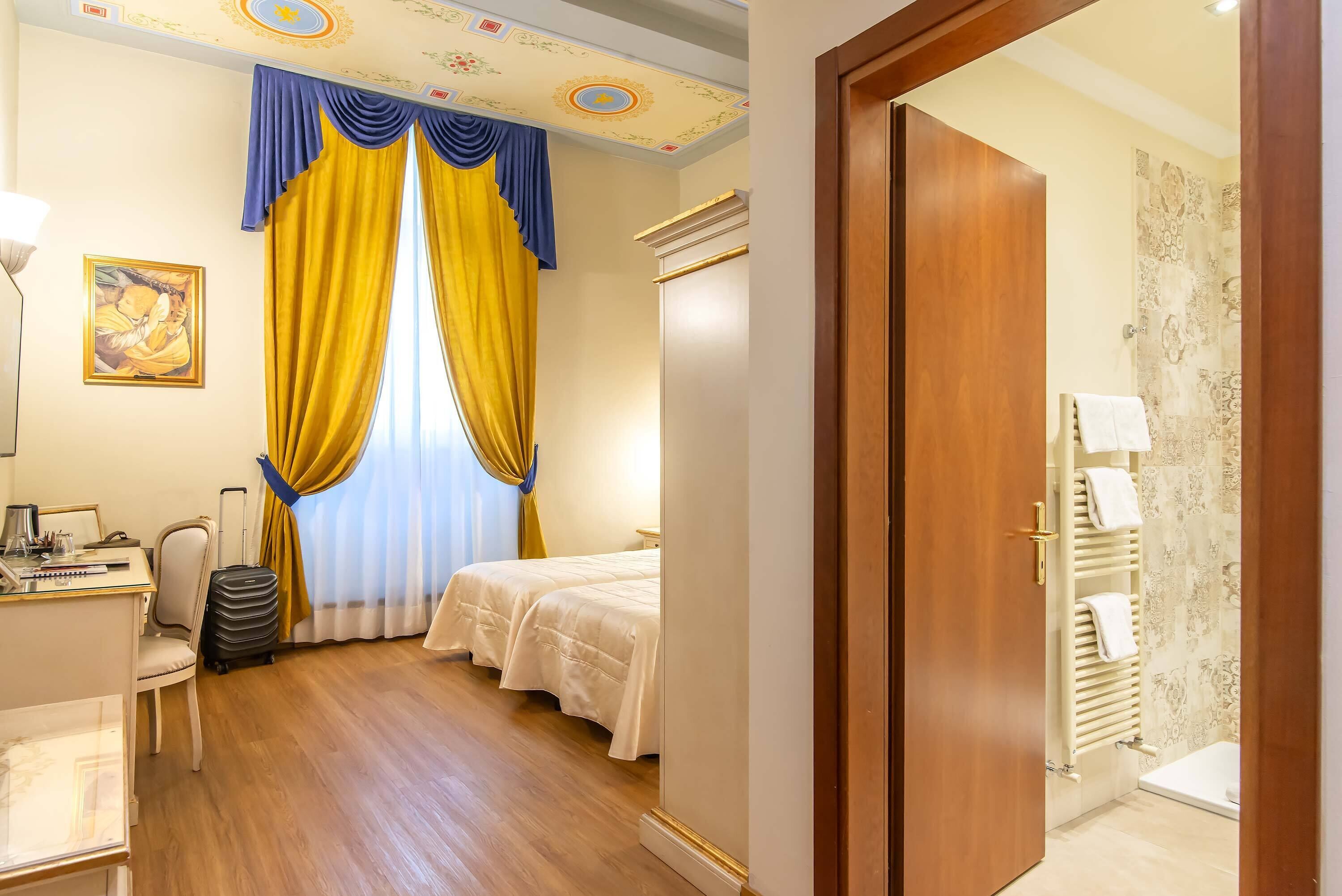 A hotel room with a bed, a desk with a chair, a suitcase, and a bathroom with a shower and towel rack.