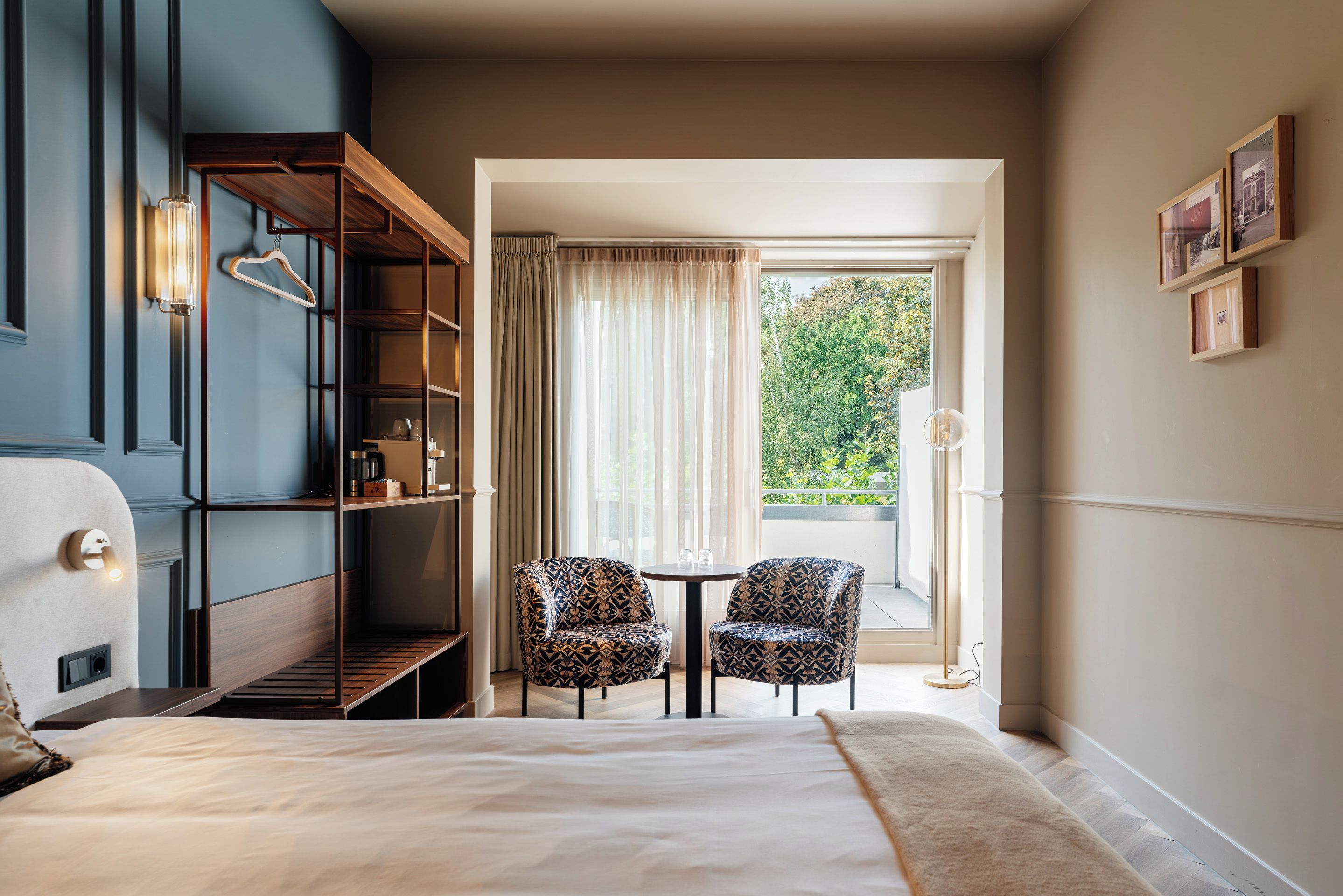 Quarto de hotel com cama, área de estar com duas cadeiras, estante de livros e janela com cortinas.