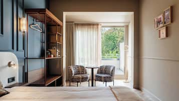 Quarto de hotel com cama, área de estar com duas cadeiras, estante de livros e janela com cortinas.