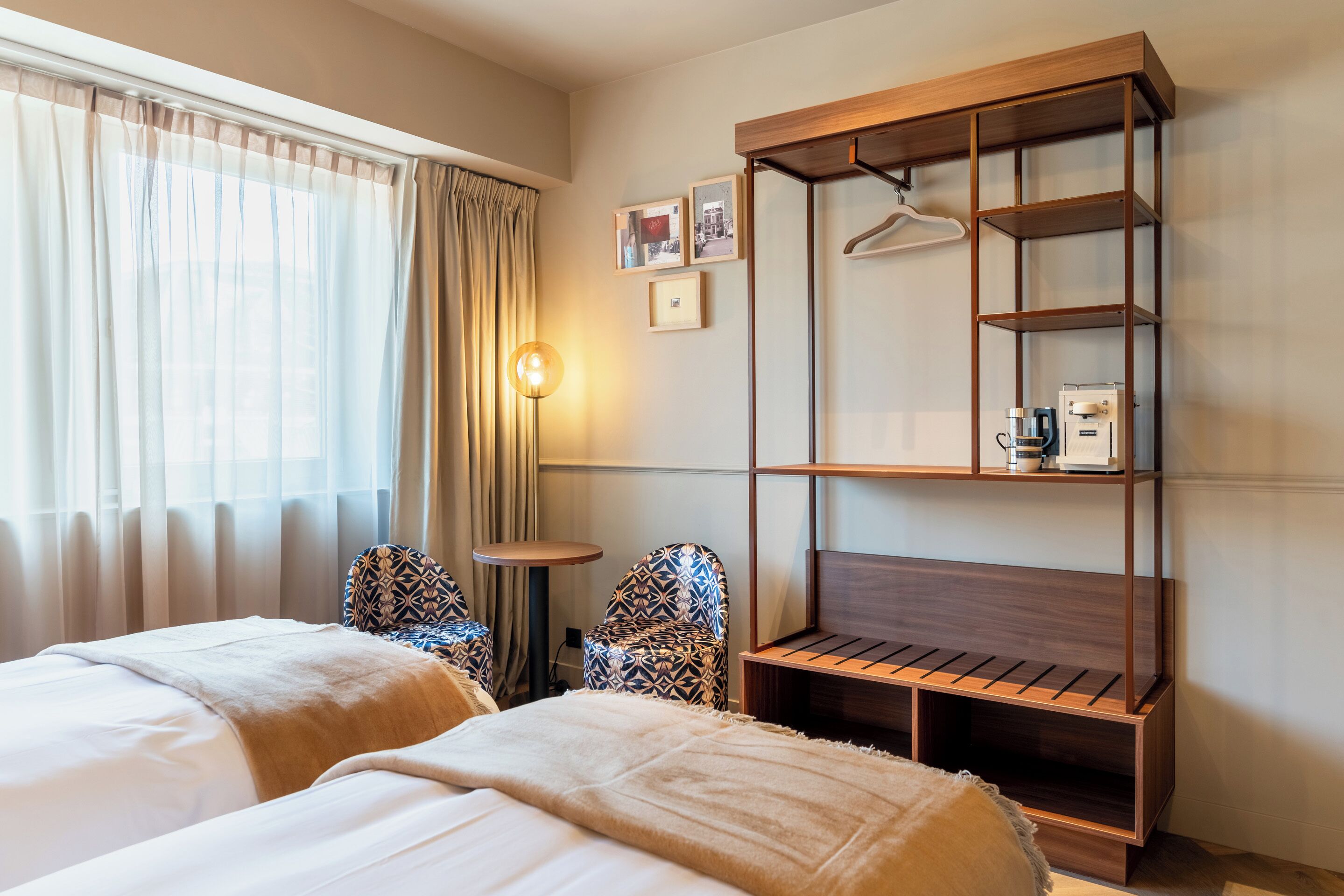 Une chambre d’hôtel avec deux lits, une petite table, des chaises à motifs, une étagère en bois avec un porte-manteau suspendu et une lampe.