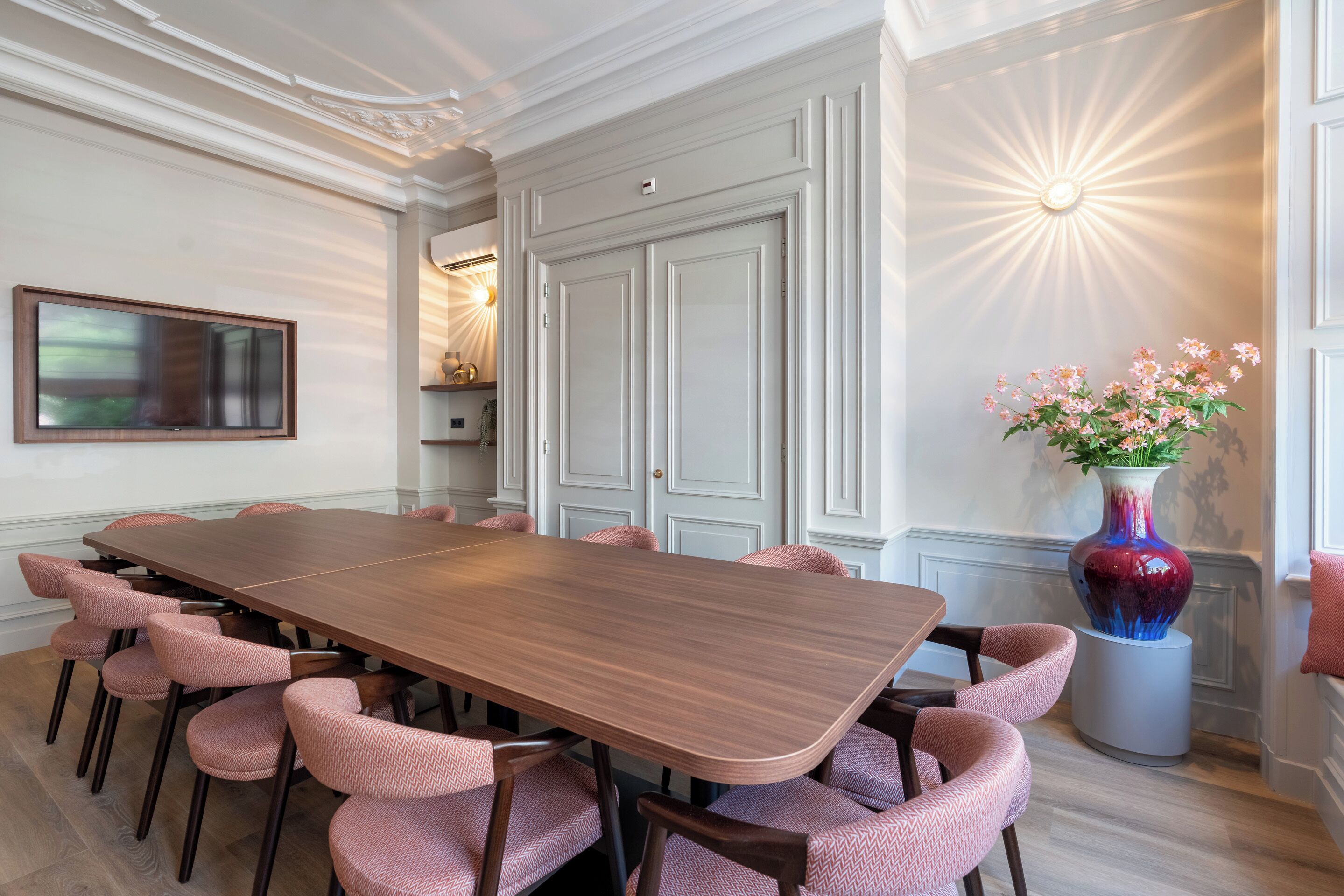 A conference room with a long wooden table, pink upholstered chairs, a large vase with flowers, and a wall-mounted TV.