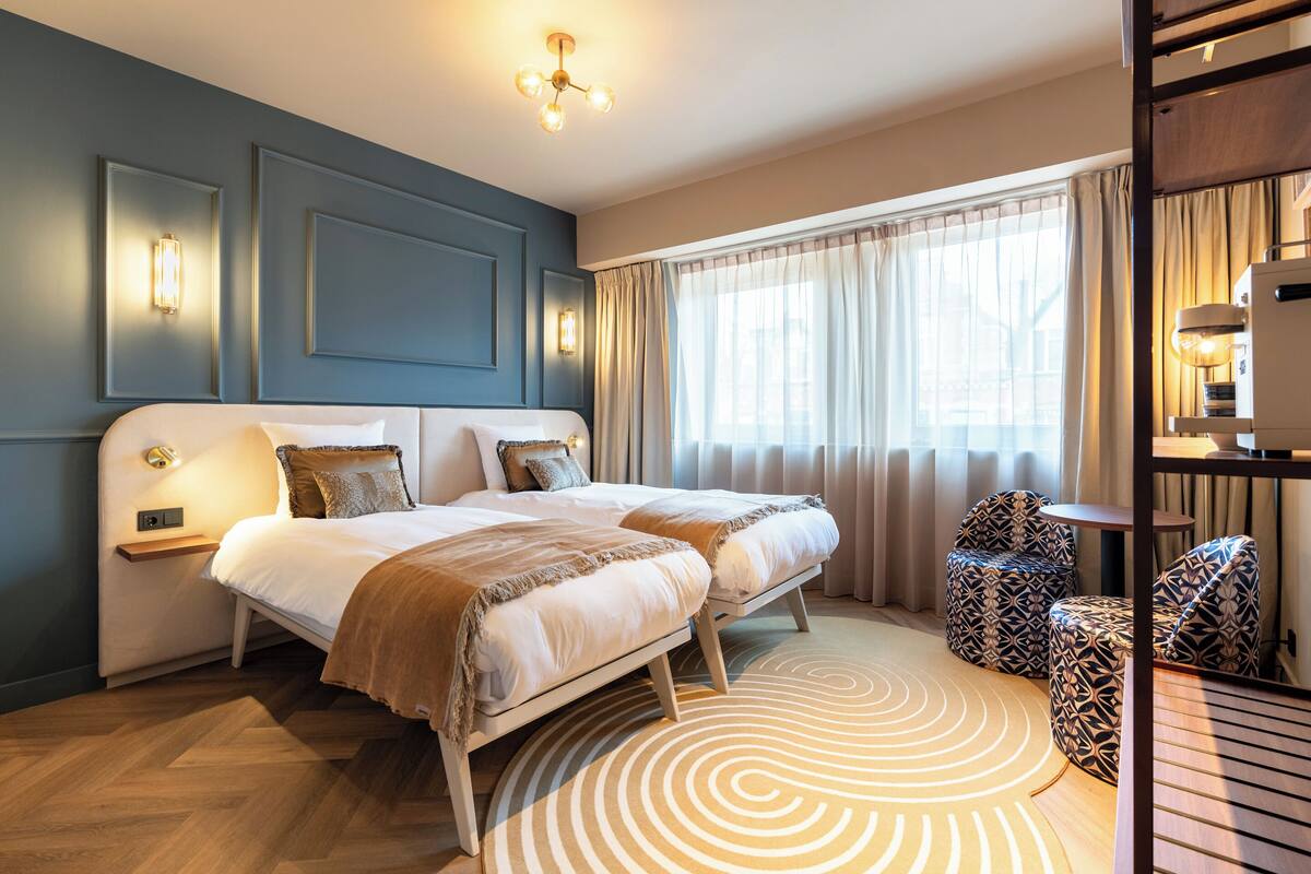 A hotel room with a double bed, two bedside tables, a patterned rug, and a view of the outdoors through sheer curtains.