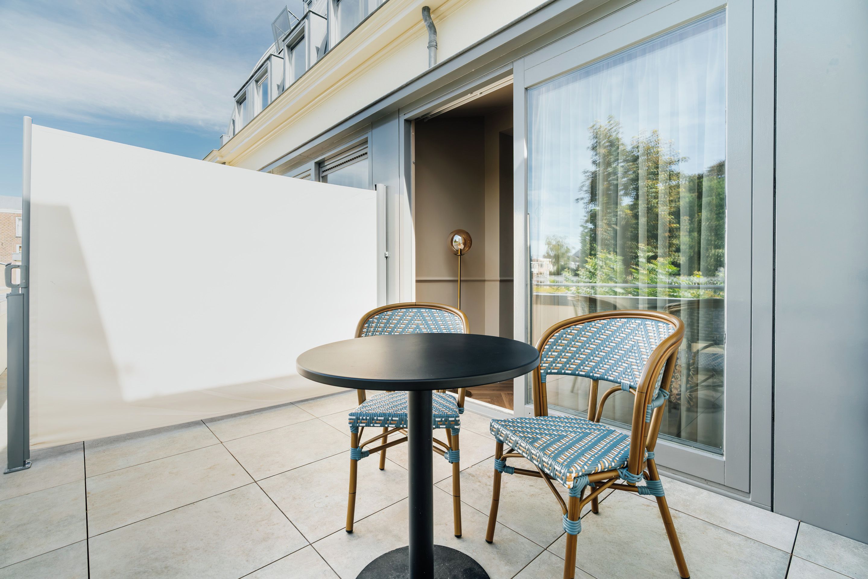A balcony with two wicker chairs and a round table, a sliding glass door, and a view of trees and buildings.