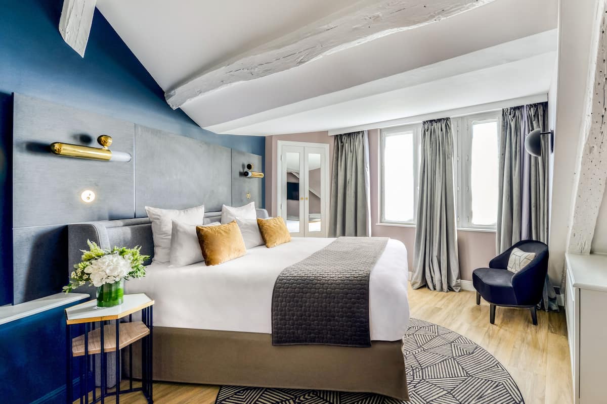 A modern hotel room with a large bed, a bedside table with a vase of flowers, a chair, and a view of the city through large windows.