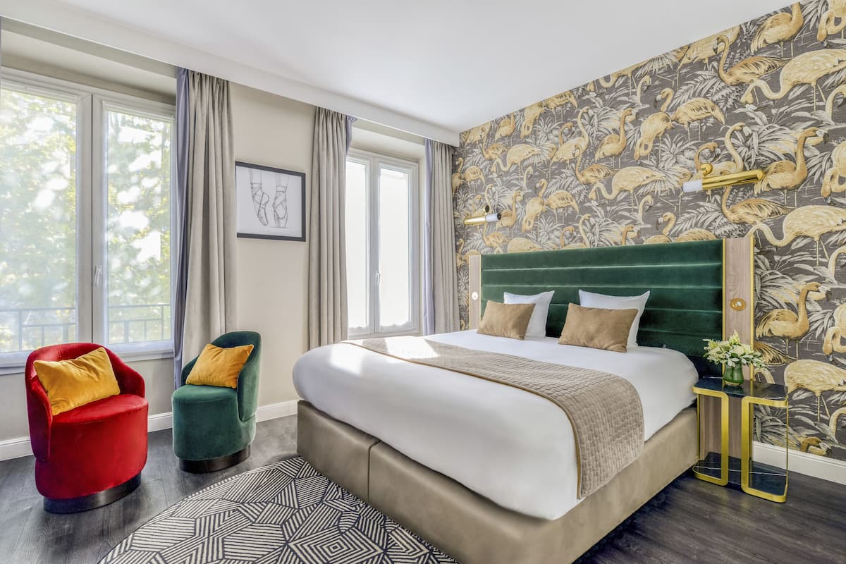A modern bedroom with a large bed, two armchairs, a patterned wallpaper, and a view of trees through the windows.