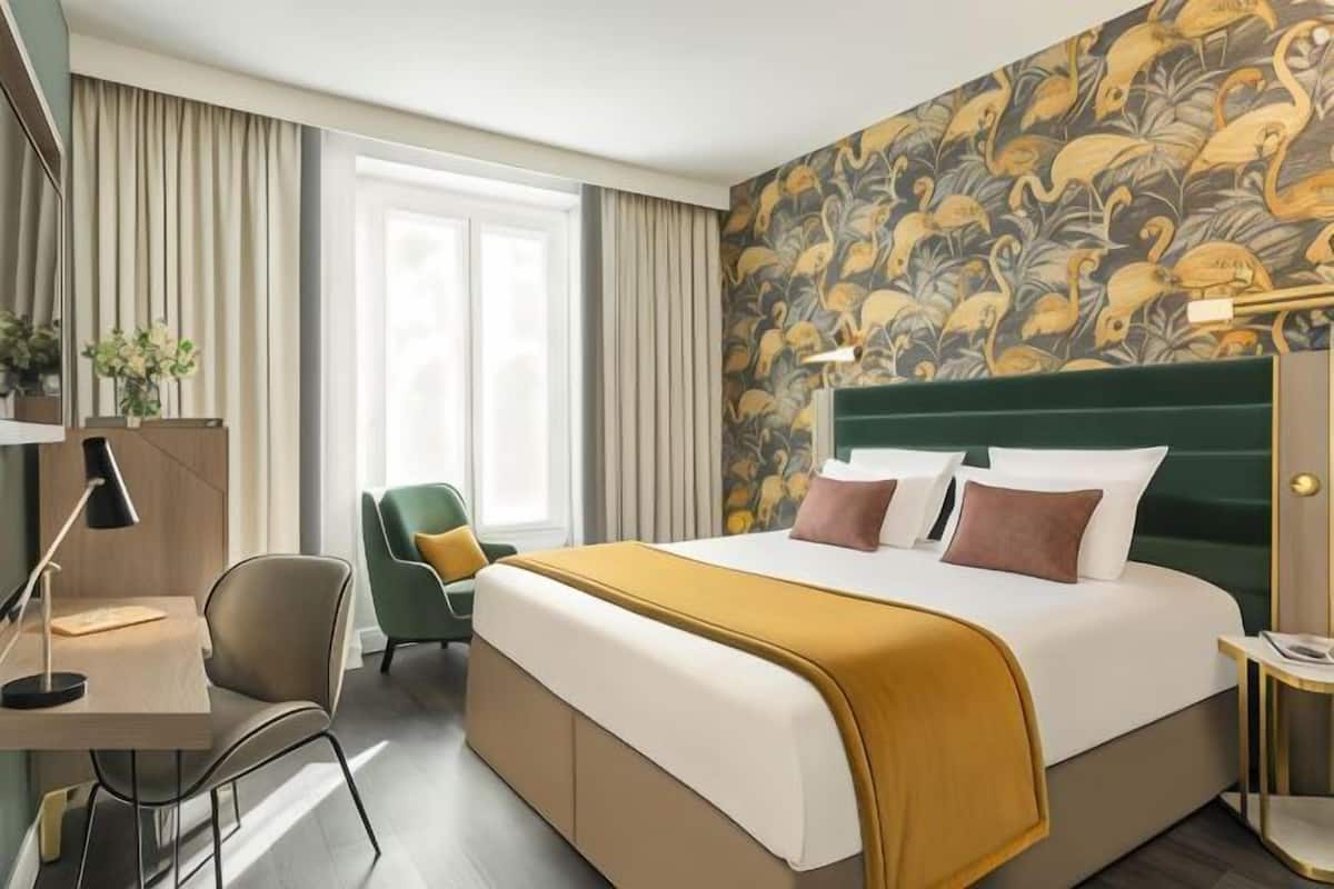 A hotel room with a large bed, a desk with a lamp, a chair, and a window with curtains.