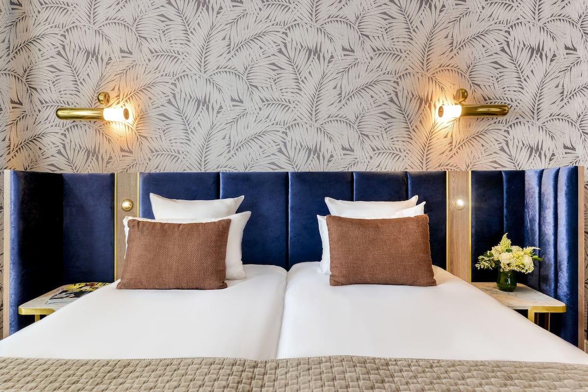 A double bed with a blue headboard, two brown pillows, and a white bedspread. There are two wall sconces with lights on, and a small table with a vase of flowers to the right.