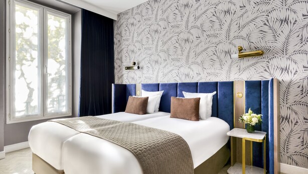 A double bed with a beige quilt, a blue headboard, and a small bedside table with a vase of flowers. - Hotel Opéra Opal (Paris)