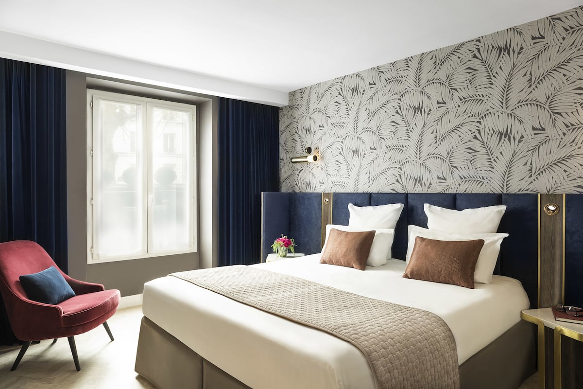 A hotel room with a large bed, a red armchair, a nightstand with a lamp, and a window with curtains.