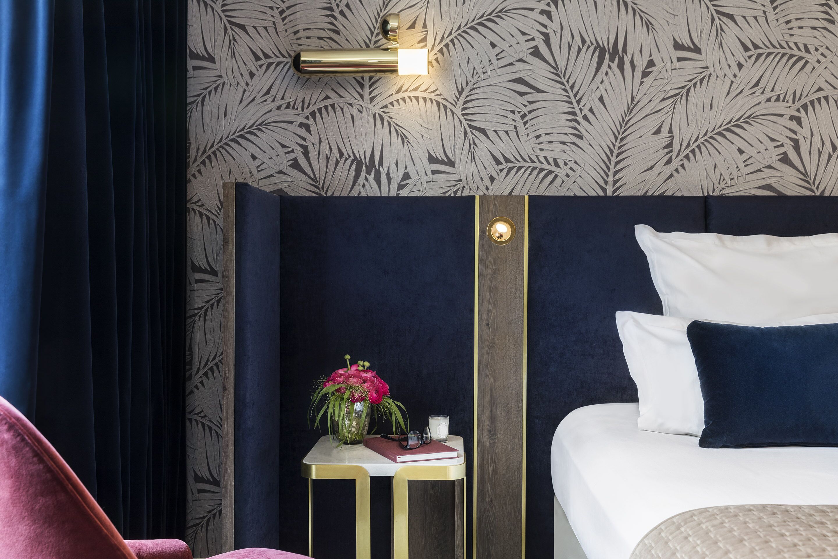 A hotel room with a dark blue headboard, a small bedside table with a vase of flowers, a book, and a cup.
