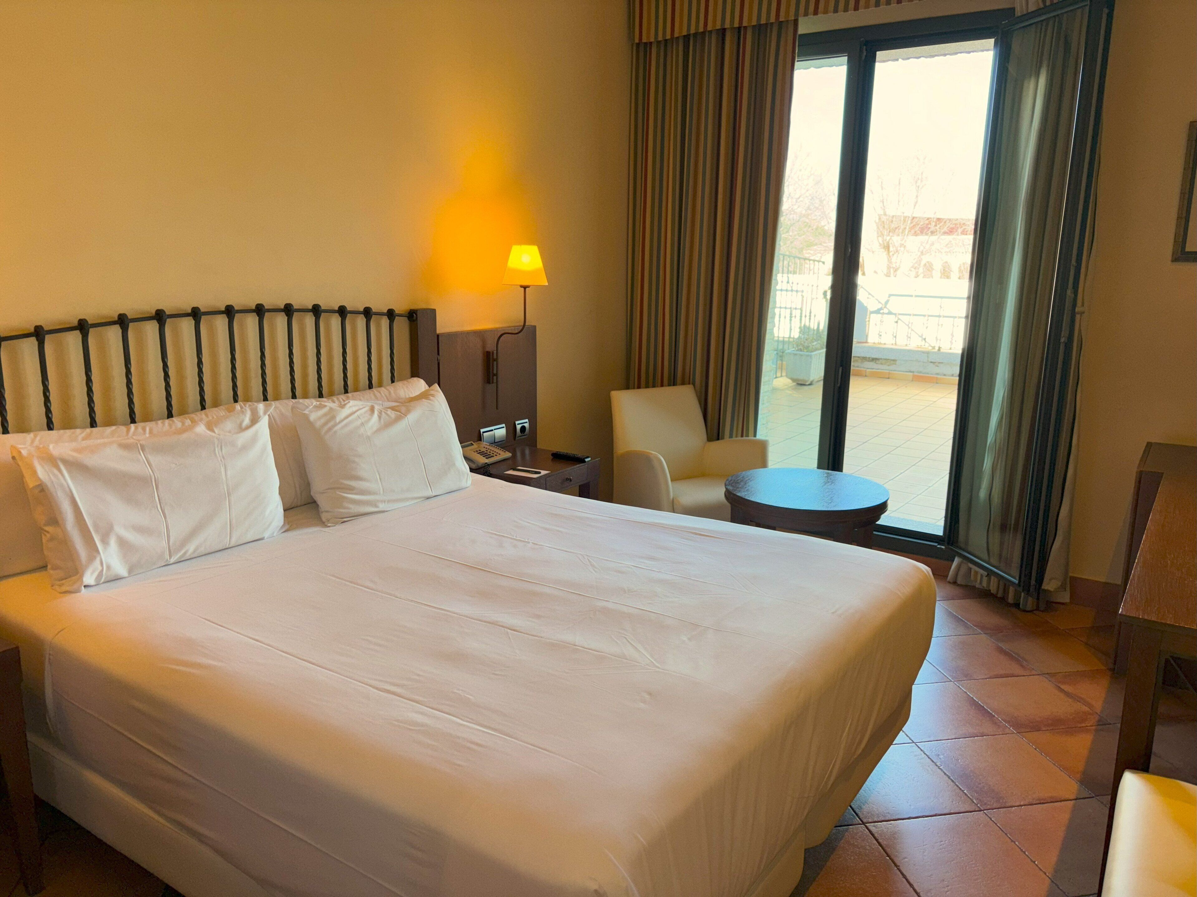 Habitación de hotel con una cama grande, una mesita de noche con lámpara, una silla y vistas al exterior a través de una puerta corredera.
