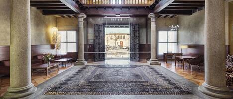 A grand lobby with a central open space, stone columns, a balcony, and a view of the street.